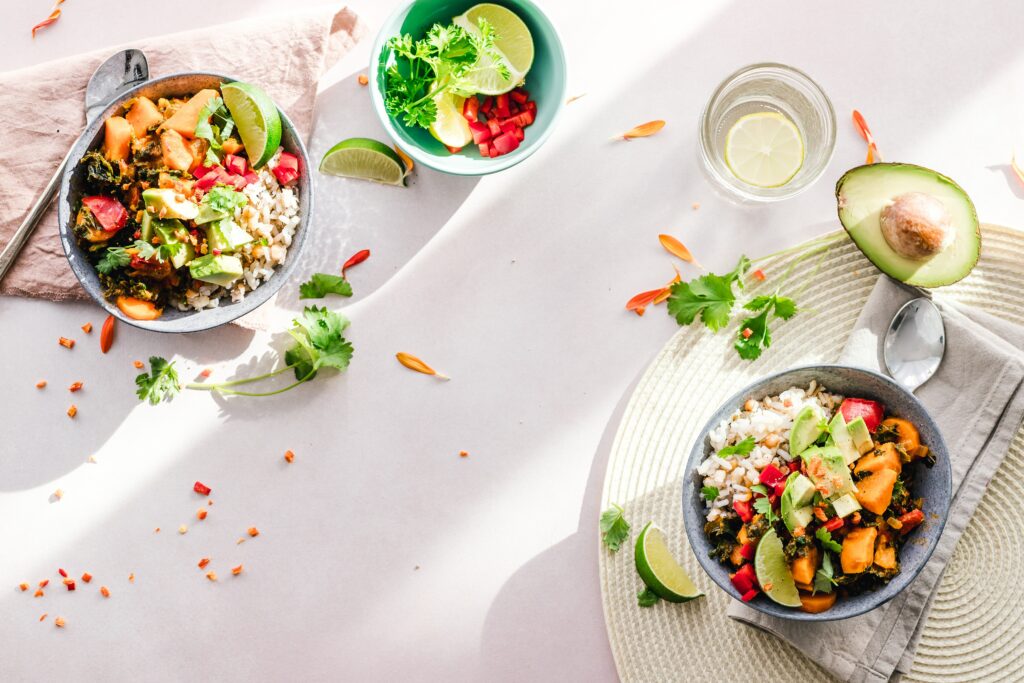 ein Tisch mit weißer Tischdecke und ein paar Schalen mit gesundem Essen, Salat, für viel Vitamine, Ballaststoffe und Proteine, mit Limette, Avocado, Kürbis, Hülsenfrüchten, ein Glas mit Wasser und Zitrone weil viel trinken wichtig ist